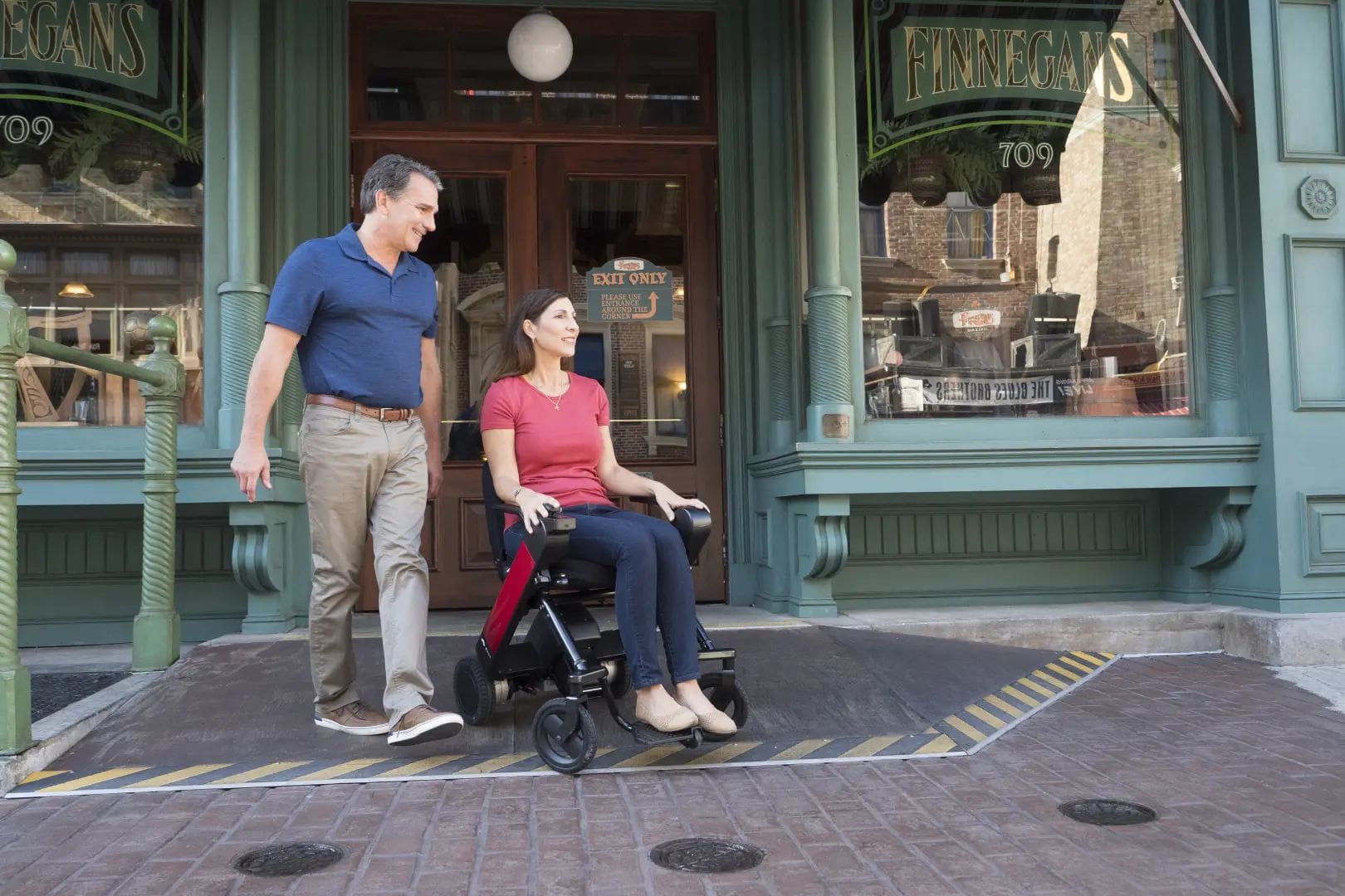 A man and woman standing in front of a building in a wheelchair.