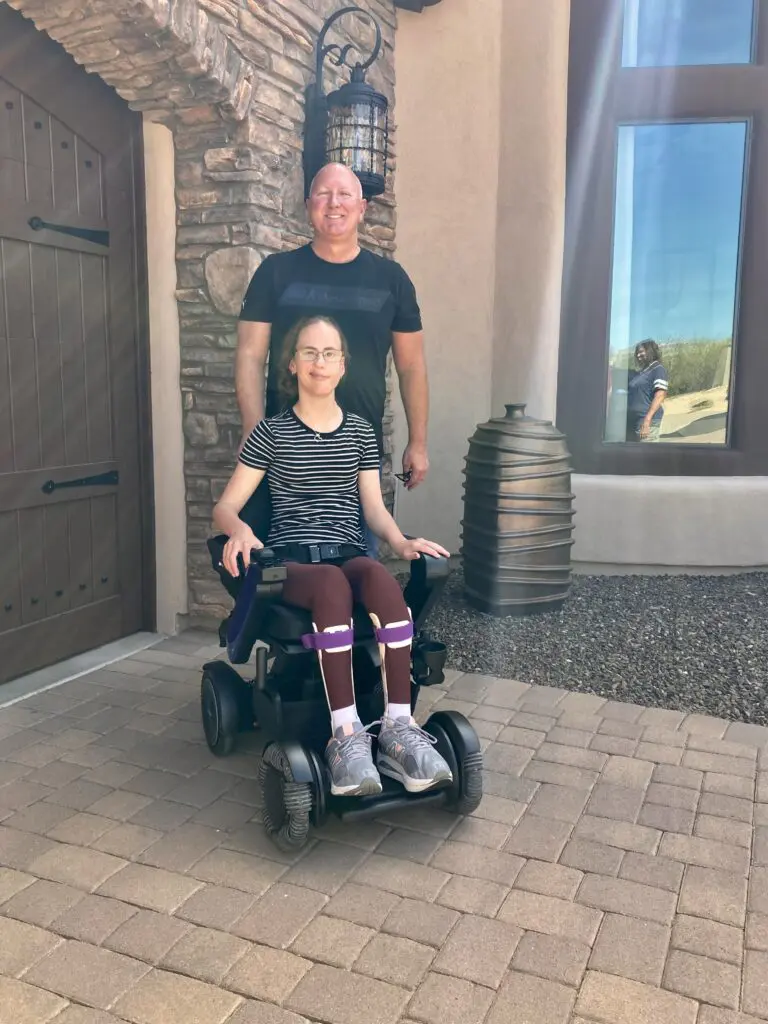 A man and woman standing in front of a house in a wheelchair.