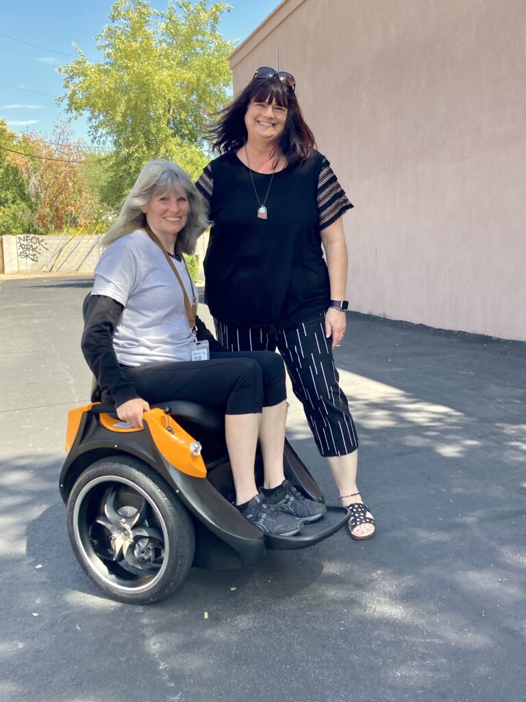 Two women standing next to a wheel chair in a parking lot.
