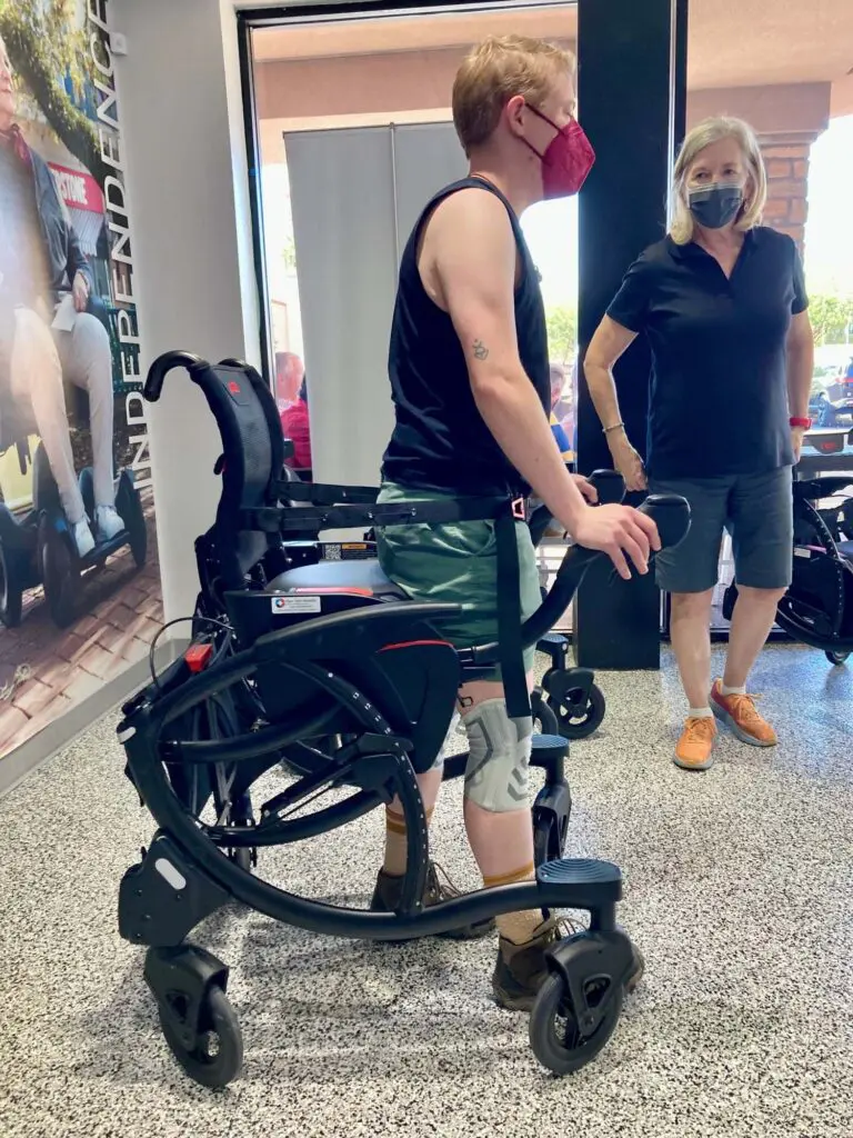 A man wearing a mask is standing next to a person in a wheelchair.