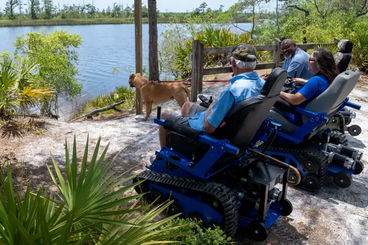 A group of people sitting on power wheelchairs near a lake.